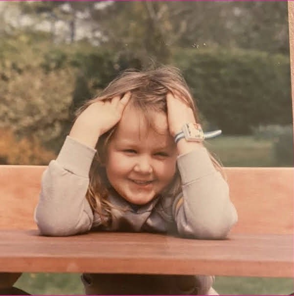 A photo of a five year old Kat, owner of Letters from Poppy. Kat is sat on a bench, leaning forward with her elbows on a matching table. Her hands are in her messy brown hair. She is looking away from the camera and smiling cheekily.